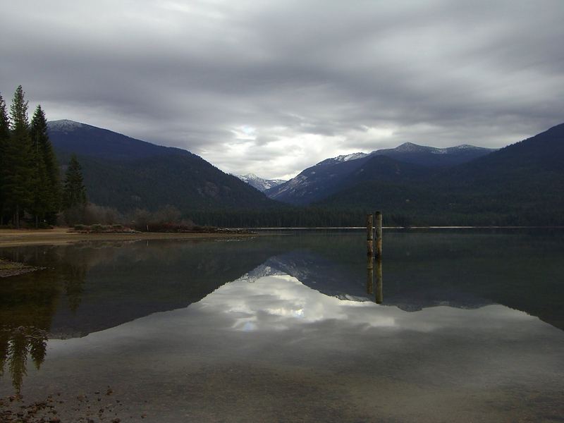 Still water on Priest Lake reflecting forested mountains under a gray autumn sky