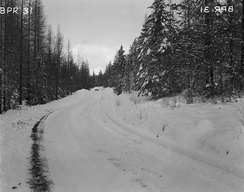 Black-and-white photo of a plowed road between snowbanks and evergreens, 1961